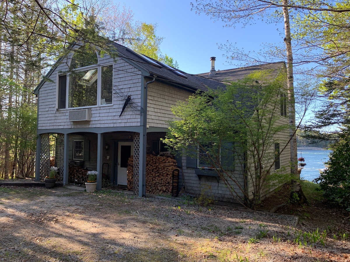The exterior of the waterfront home is framed by lush greenery, showcasing a shingled facade. Woodpile storage is neatly arranged on one side. A large window offers a glimpse of the water, while a tranquil path leads to the entrance.