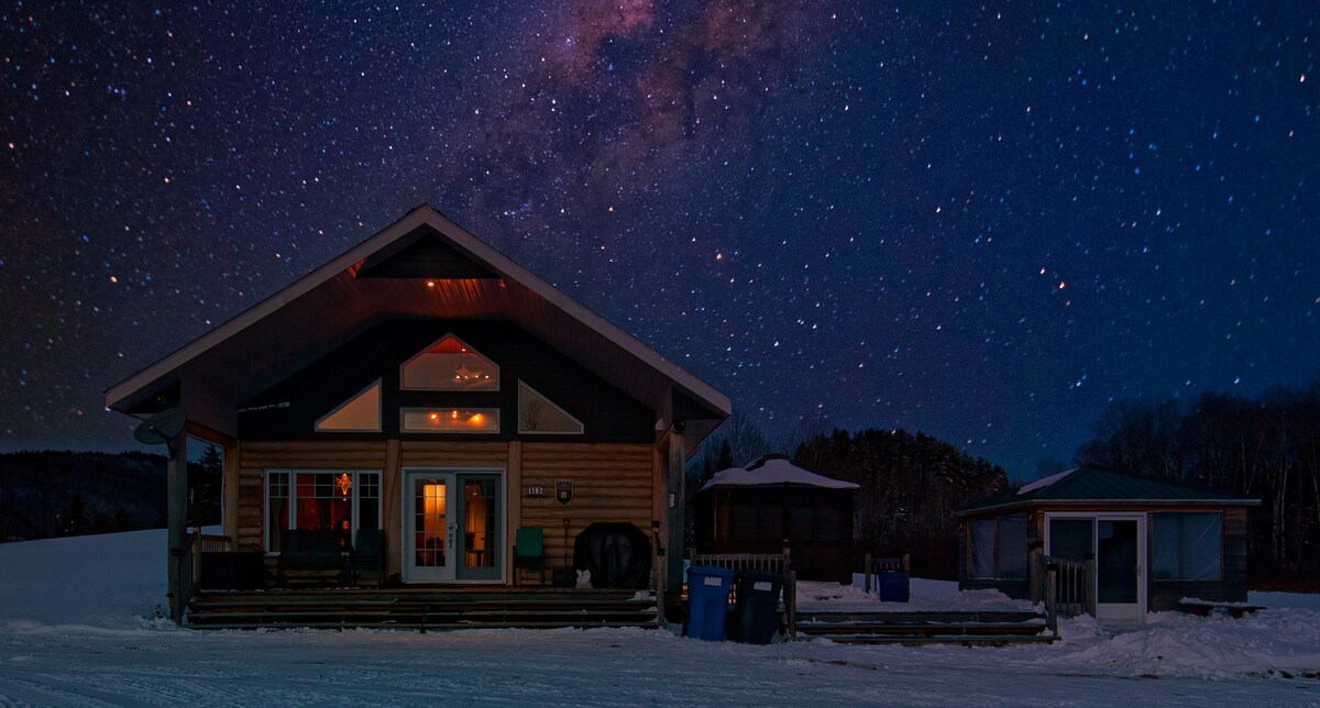 A spacious chalet is depicted against a starry night sky, showcasing its large front windows and inviting porch. The surrounding snow-covered landscape adds to the serene atmosphere, while distant trees enhance the natural setting.