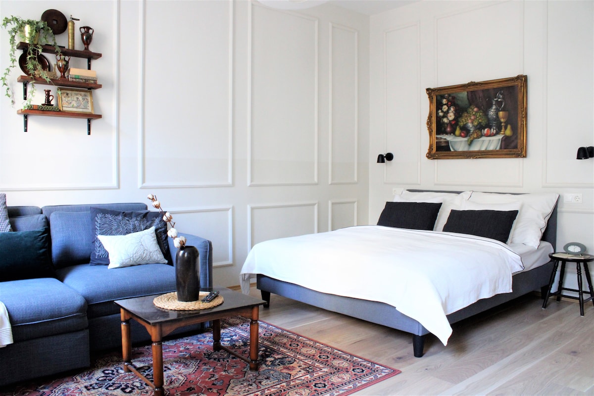 A cozy living space features a comfortable sofa with decorative pillows and a spacious bed dressed in white linens. A small side table rests beside the bed, and a colorful area rug adds warmth to the wooden floor. Wall sconces provide subtle illumination, enhancing the inviting atmosphere.