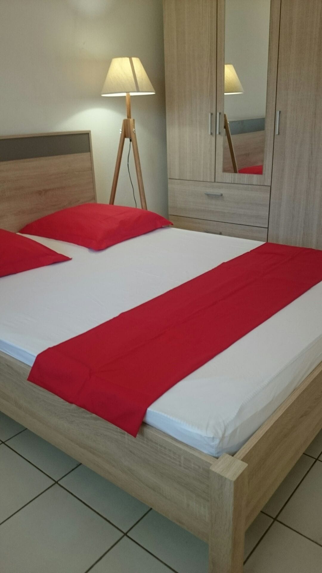 A well-lit bedroom features a wooden bed frame with a white mattress adorned with red pillows and a matching bed runner. A floor lamp provides additional light, while mirrored cabinets offer storage space, contributing to a clean and organized appearance.