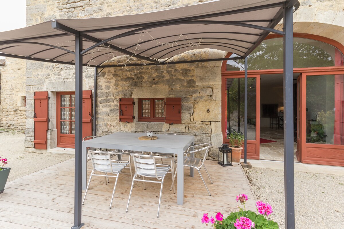 A wooden terrace features a gray table surrounded by four metal chairs under a fabric canopy. Red window shutters and stone walls provide a rustic backdrop, while potted flowers add a touch of color to the space.