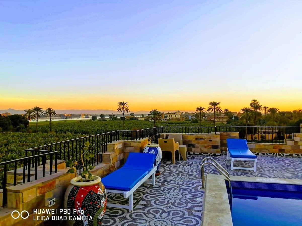 A rooftop terrace features a swimming pool beside blue sun loungers, offering sweeping views of the Nile and surrounding palm trees under a vibrant sunset sky. The terrace boasts intricate tile patterns, enhancing the overall scenic atmosphere.