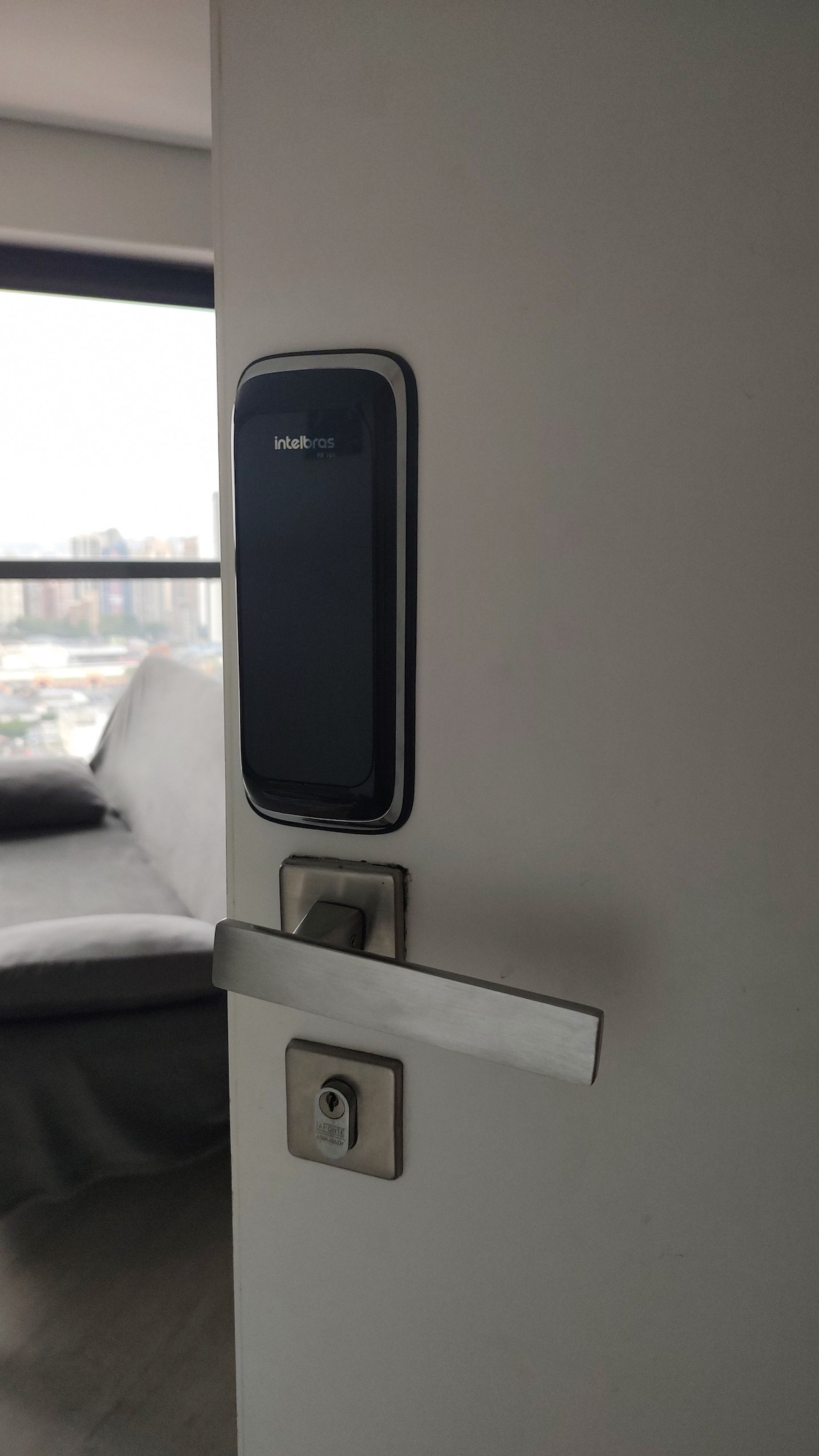 A modern door lock system is displayed on a white door, featuring a sleek, black panel. The stainless steel handle complements the contemporary design. In the background, a blurred view of a cozy seating area is visible, offering hints of natural light entering through nearby windows.