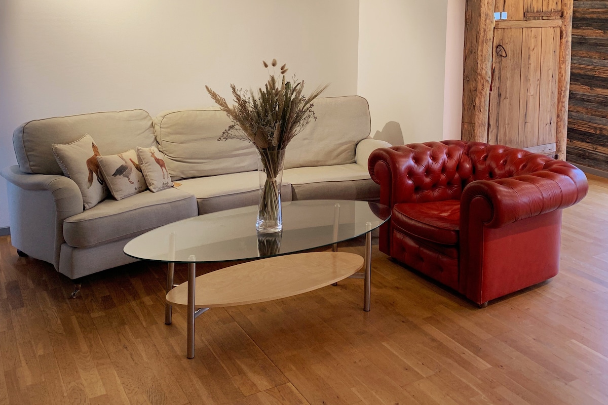 A light and airy living area is presented with a beige sofa and a red leather armchair, creating a comfortable seating arrangement. A glass coffee table sits in the center, topped with a vase containing dried floral elements. Soft wooden flooring completes the inviting setup.