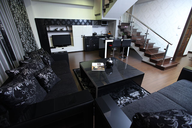 A stylish living area features a dark color scheme with plush seating and decorative pillows. A glass coffee table is centrally placed, while a sleek kitchen area is visible in the background, equipped with modern appliances. A staircase leads to the upper level.