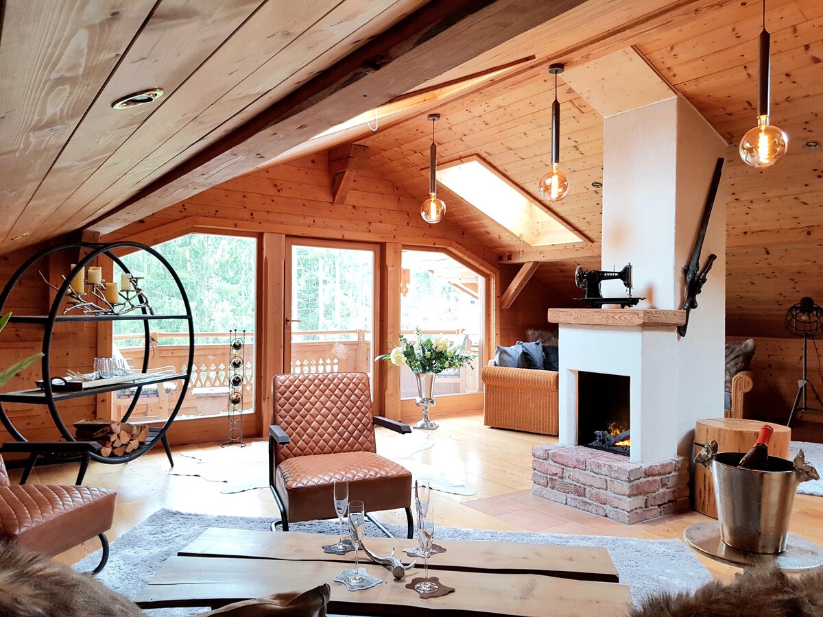 A spacious lounge area features wooden walls and ceilings, with natural light flooding in from multiple windows. A cozy fireplace is set against a brick backdrop, while stylish seating arrangements include two textured chairs and a wooden coffee table. Decorative elements enhance the inviting space.