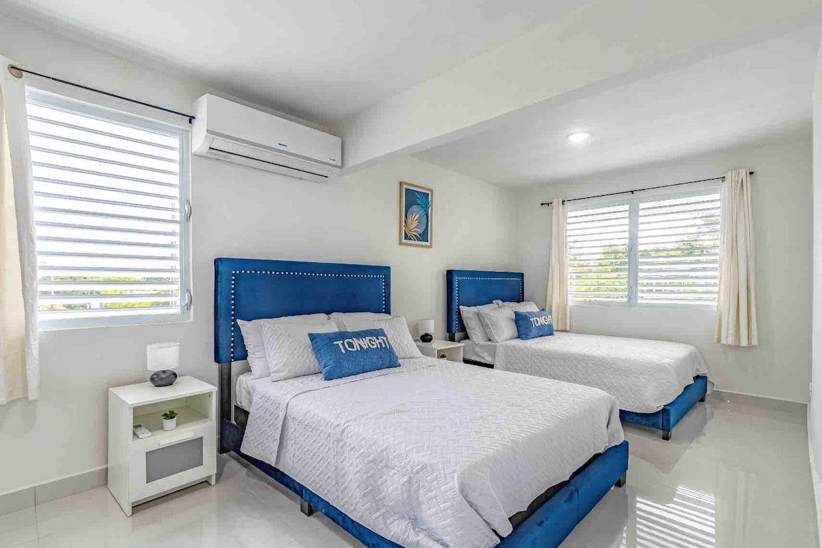 A bright room is furnished with two spacious beds, each featuring blue headboards. Natural light enters through two windows with horizontal blinds. Modern amenities are highlighted by a nightstand and an air conditioning unit mounted on the wall.