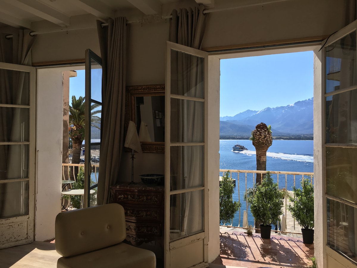 Double doors open to a stunning view of the sea and mountains, with a boat visible on the water. Potted plants line the balcony, providing a touch of greenery. Soft curtains frame the scene, blending indoor comfort with the outdoor landscape.