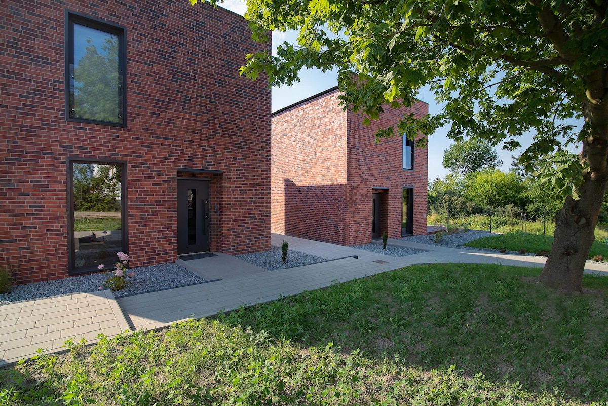 The exterior view captures two modern brick houses with large windows framed in black. A paved path leads to the entrances, surrounded by well-maintained grass and shrubs. A tree provides shade to the area, enhancing the tranquil setting in the countryside.