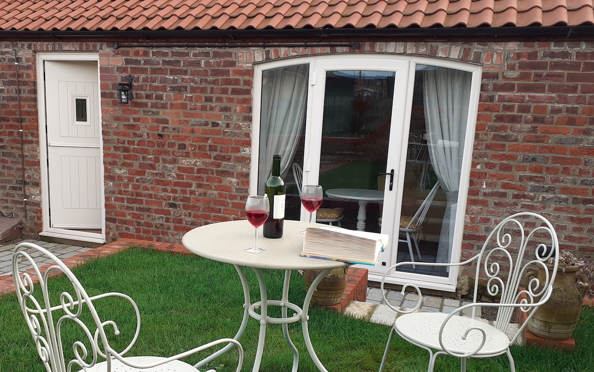 An outdoor seating area features a round table set with two glasses of red wine and a book resting on the surface. Three white wrought iron chairs surround the table, while a glass door provides access to the interior of the barn conversion, framed by curtains.