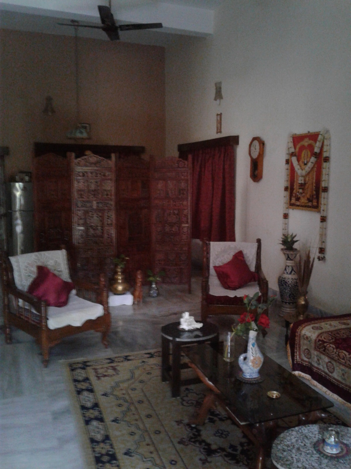 A calming living area is shown, featuring two wooden chairs adorned with red cushions. A low coffee table sits in the center, accompanied by decorative items and fresh flowers. An intricately carved wooden divider adds charm, while wall decorations enhance the serene environment.