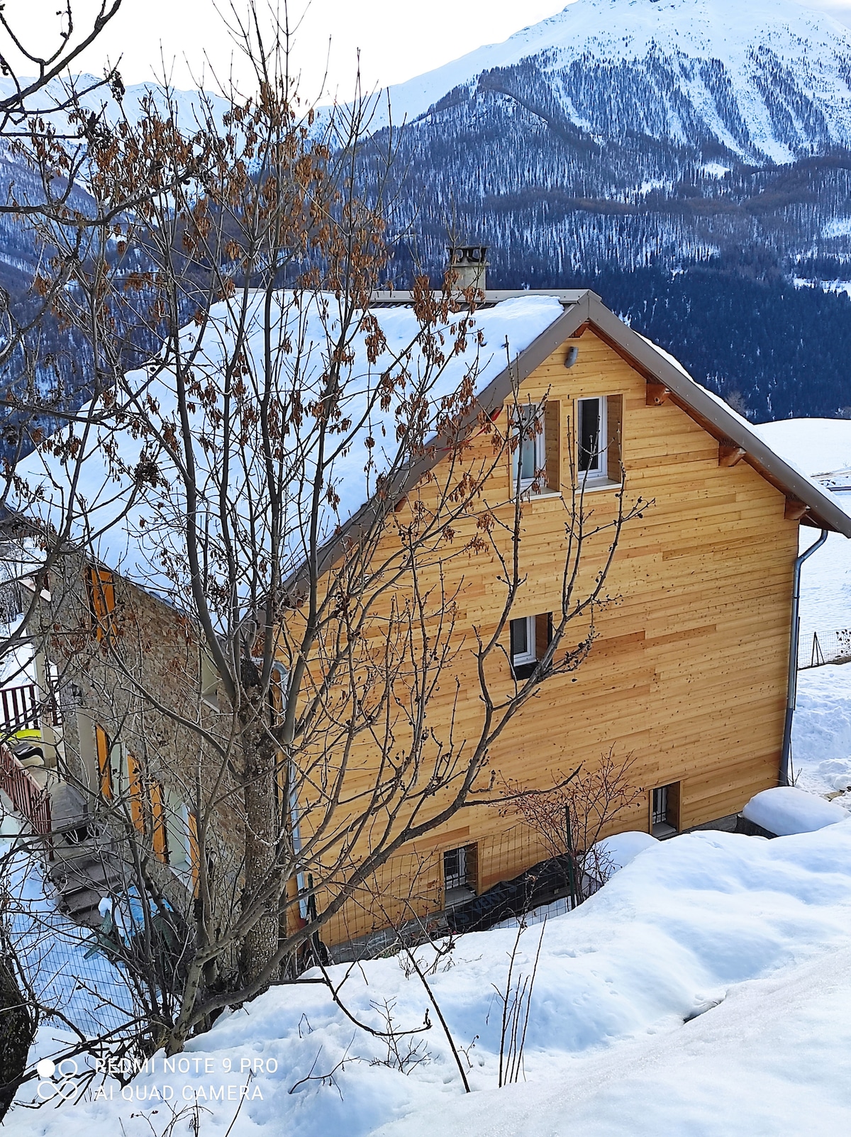 The chalet features a wooden exterior with large windows, nestled against a backdrop of snow-covered mountains. Surrounding the house are trees without leaves, indicative of winter. A layer of snow blankets the ground, enhancing the serene, alpine environment.