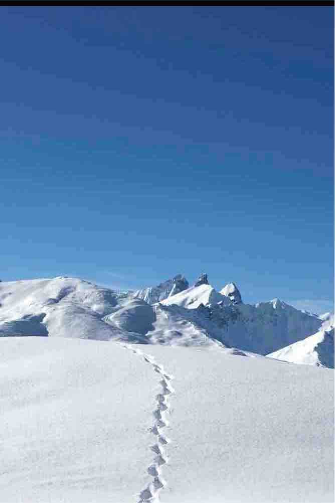 A snow-covered landscape extends under a clear blue sky, with footprints leading through the pristine snow. The distant mountains peak above the white terrain, providing a serene backdrop of rugged natural beauty.
