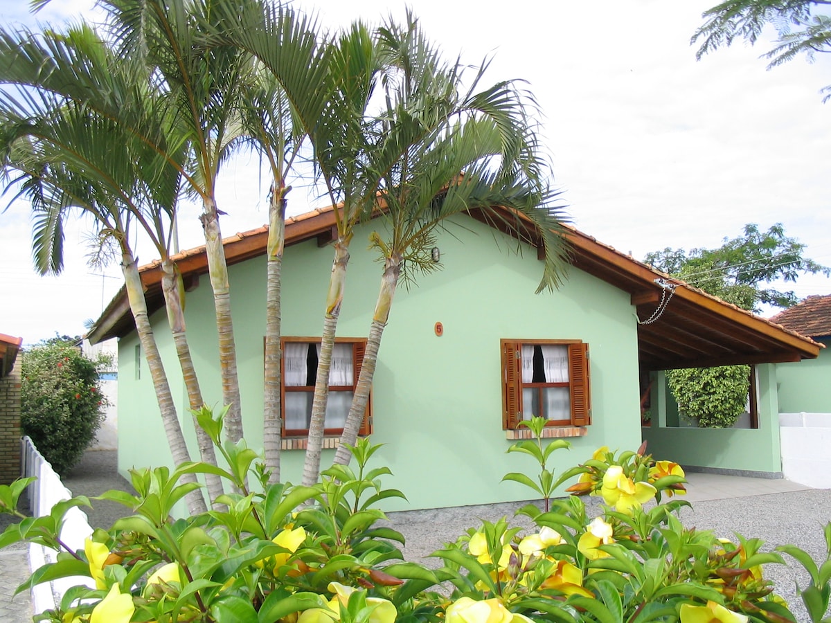 A single-story house with a light green exterior and brown wooden shutters is surrounded by tropical plants. Palm trees provide shade in the front yard, where yellow flowers add a splash of color. The roof features wooden beams that complement the home's charming design.