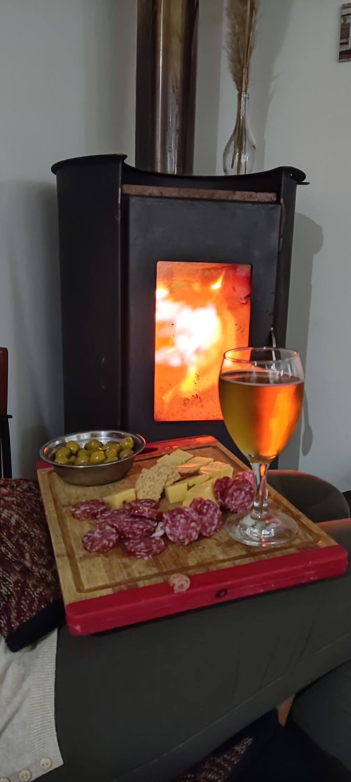 A wooden serving board displays assorted salami slices and cheese, accompanied by a bowl of olives. A glass of amber beverage rests nearby. The background features a wood-burning stove, with warm flames visible through the glass front, creating a cozy atmosphere.