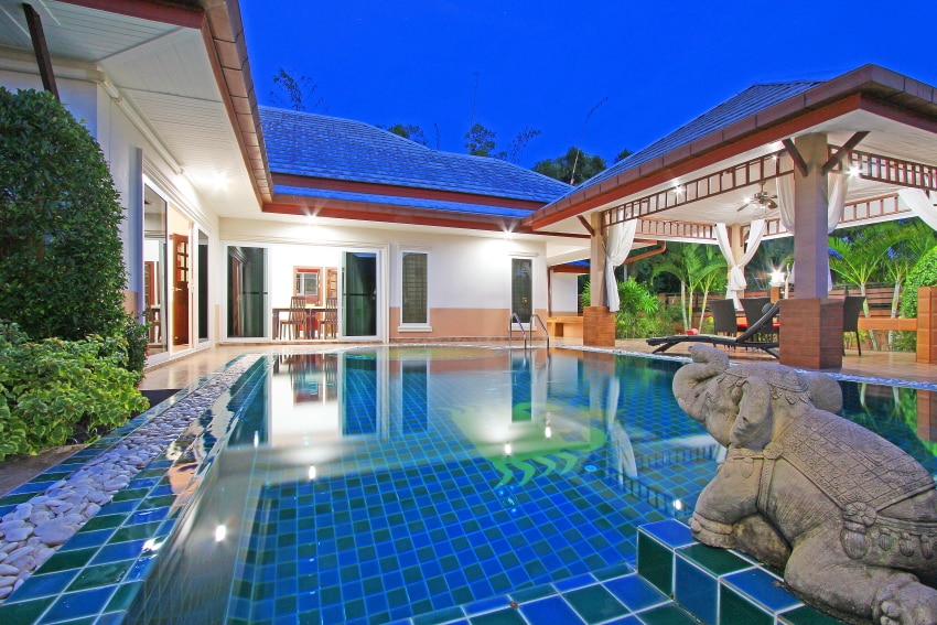 A serene swimming pool, featuring tiles in calming blue hues, is surrounded by a tropical landscape. Spacious pavilions and the villa structure are visible in the background, showcasing an inviting outdoor area perfect for relaxation.