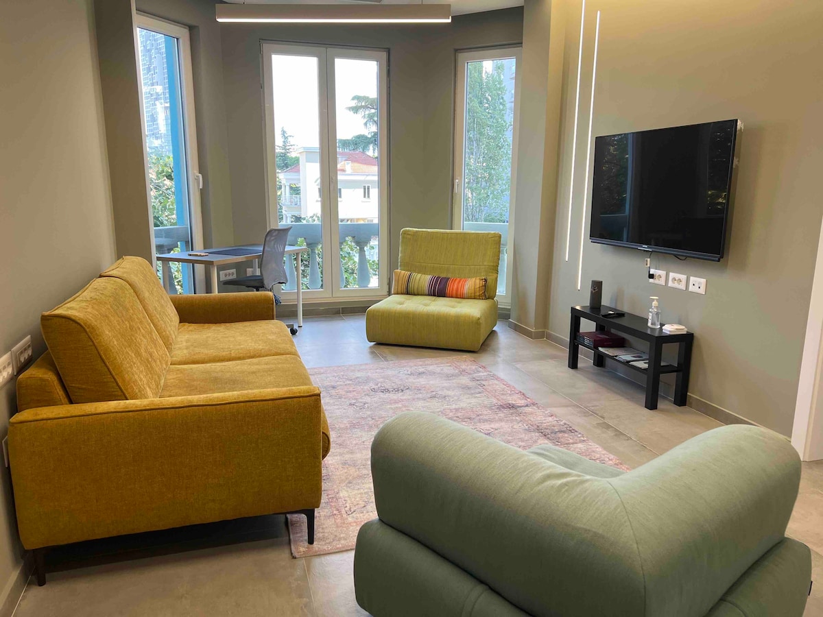 The living area is furnished with a modern sofa and a contemporary lounge chair, providing a comfortable space for relaxation. Large windows allow natural light to fill the room, while a subtle area rug adds warmth. A working space with a desk is visible in the corner.
