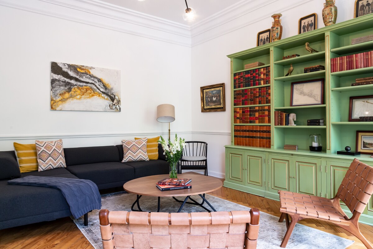 A stylish living area features a dark sectional sofa with decorative throw pillows, a round coffee table, and two leather accent chairs. The green bookshelf showcases a collection of books and decorative items. Natural light fills the space, highlighting the elegant design elements.