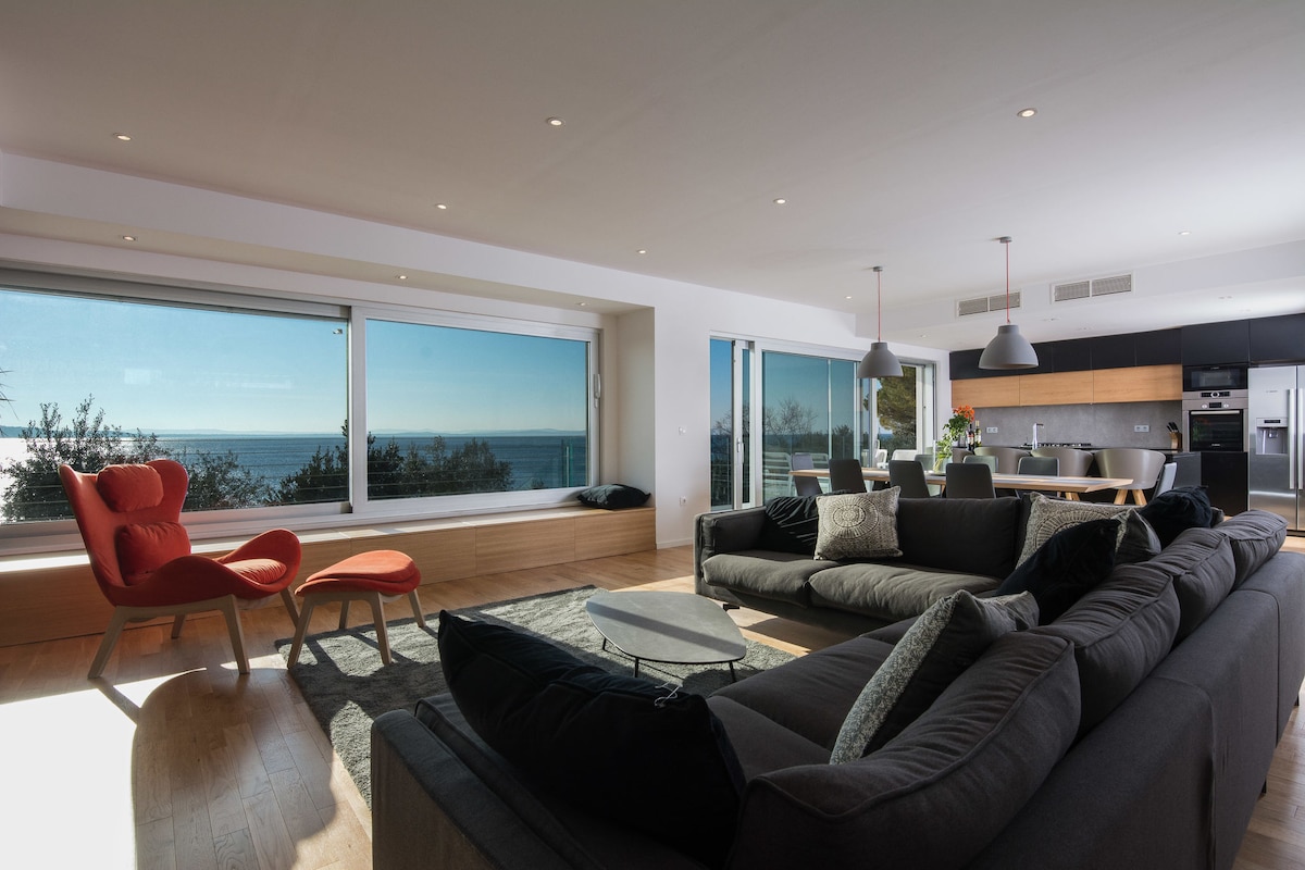 A spacious living area features large windows offering views of the sea. Comfortable seating includes a dark sectional sofa and accent chairs. A round coffee table is centered on a light-colored rug, while a dining area with modern lighting is visible in the background.