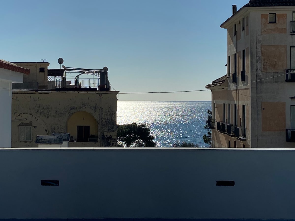A view of the shimmering ocean is captured from the balcony, with sunlight reflecting off the water. Surrounding structures, featuring balconies and textured walls, frame the scene, emphasizing the coastal setting.