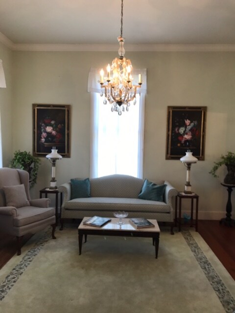 A charming sitting area features a soft blue sofa, complemented by armchairs and decorative lamps. Two floral paintings adorn the walls, and a small coffee table sits in the center. Natural light streams in through the windows, highlighting the elegant chandelier overhead.