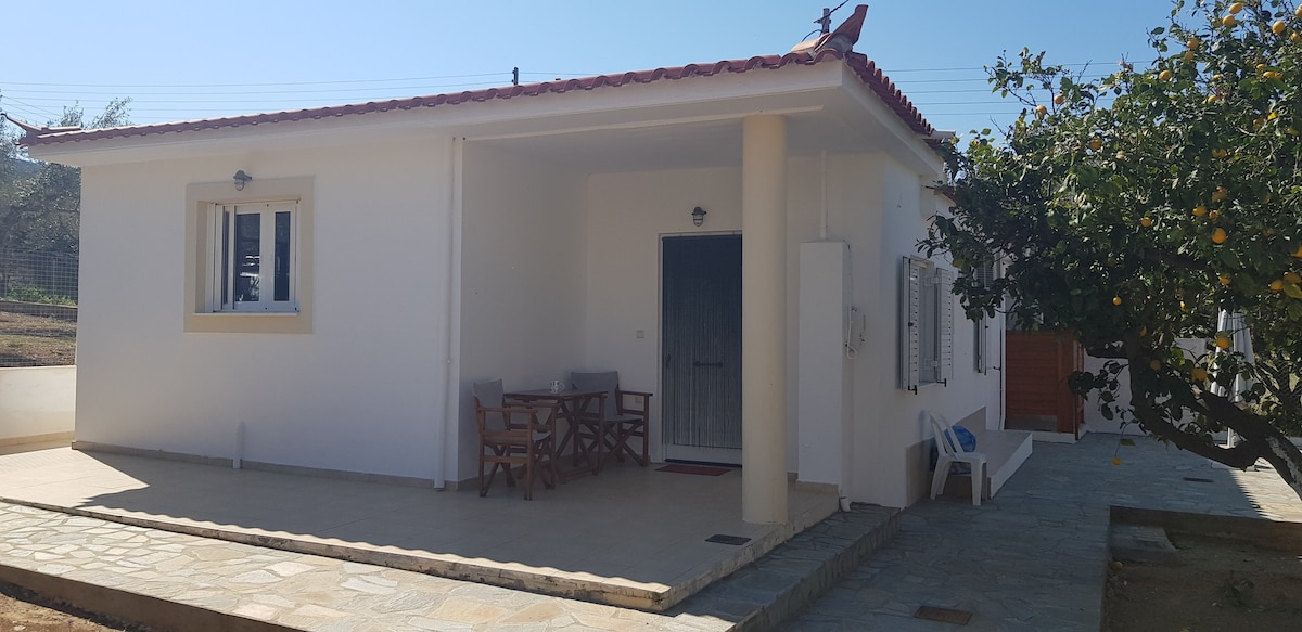 The exterior of the house is presented, featuring a white facade with a terracotta tiled roof. A shaded seating area with wood chairs and a small table is situated at the entrance. A stone path leads to the entrance, surrounded by greenery and citrus trees.