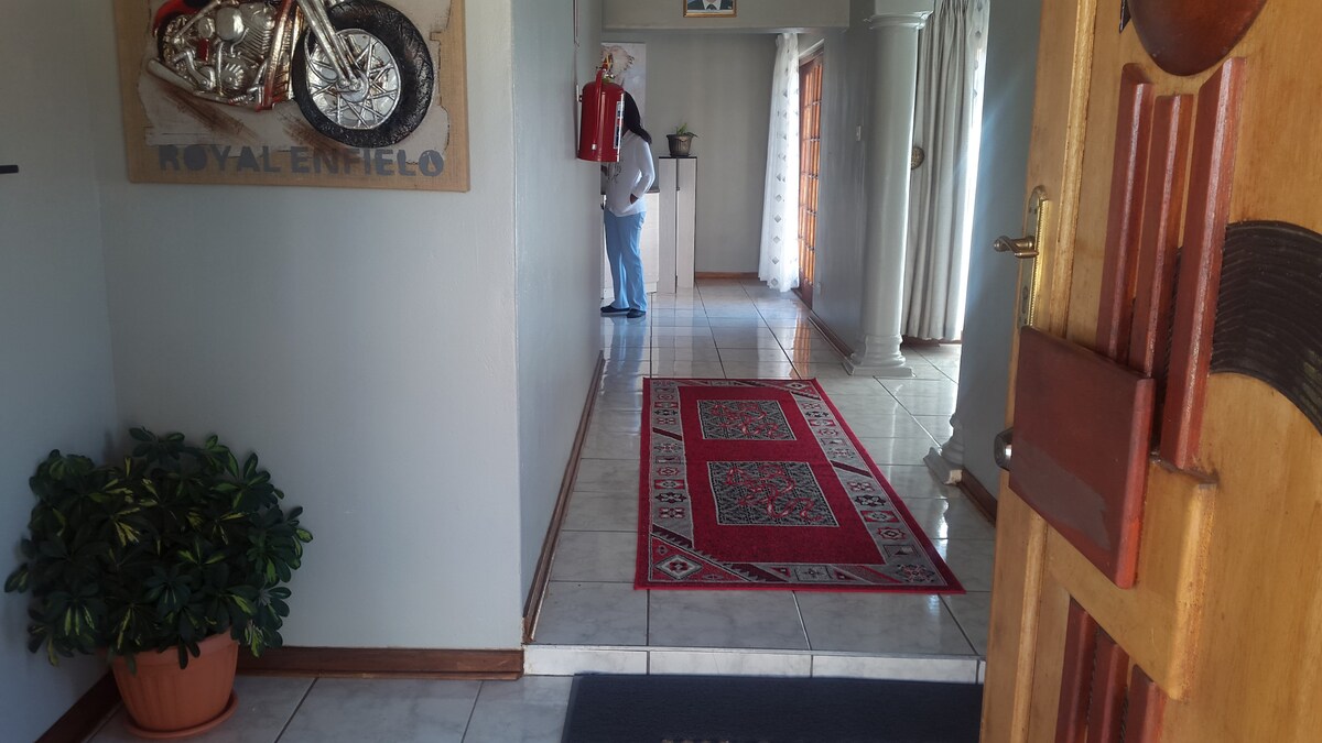 A welcoming entryway features a large area rug with intricate patterns, positioned on tiled flooring. A decorative wall art piece of a motorcycle adds character to the space. Natural light brightens the hallway, leading towards an open area with a wooden door and light-colored curtains.