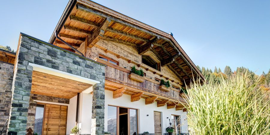 The exterior of the lodge showcases a blend of natural stone and wood materials, featuring large windows that invite light. A balcony adorned with potted greenery extends from the upper level, and the surrounding landscape complements the rustic charm of the structure.