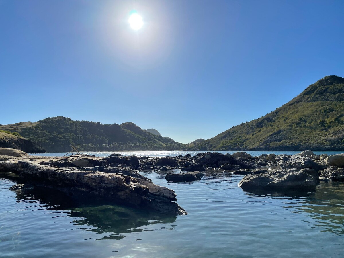 A natural landscape features calm waters reflecting a clear blue sky, with the sun shining brightly overhead. Smooth stones and logs are partially submerged, while green hills rise gently in the background, framing the serene setting.