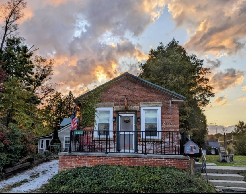 The front view of the Old Postal Cottage showcases its charming brick facade and welcoming porch. Lush greenery surrounds the exterior, and a flag can be seen displayed. A cloudy sunset sky adds a gentle glow, enhancing the inviting atmosphere of the residence.