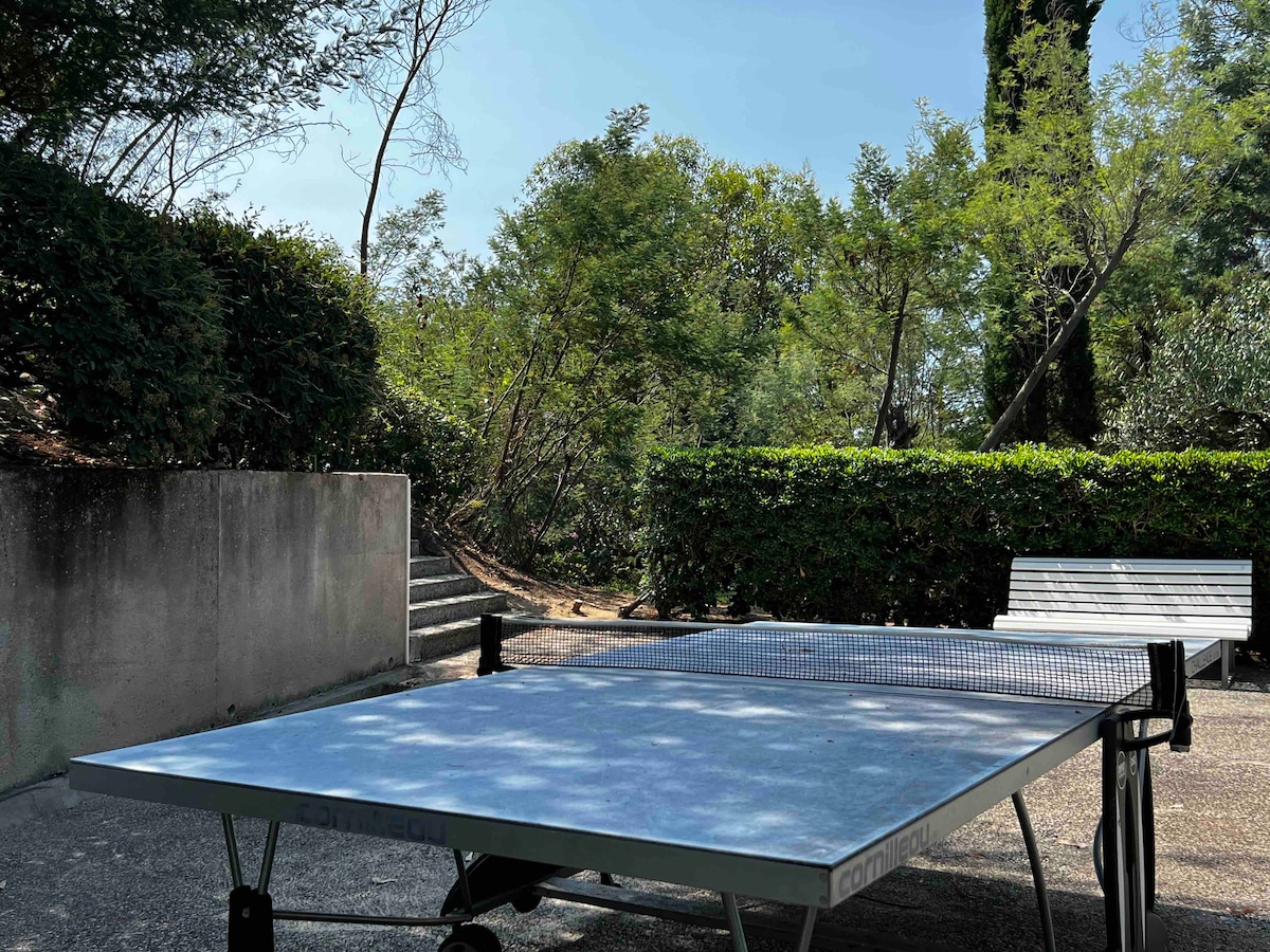 A ping-pong table is positioned outdoors, surrounded by lush greenery and shrubs. Sunlight filters through the trees, creating a bright and inviting atmosphere. A bench is visible in the background, providing a spot for relaxation amid the natural setting.