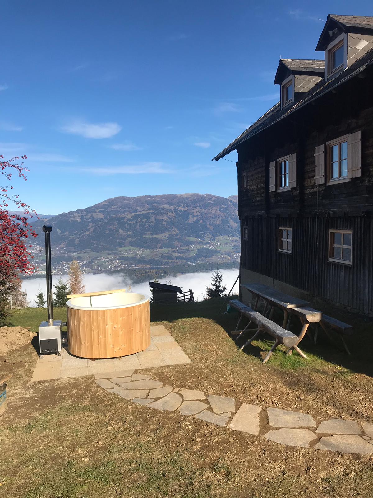 A wooden hot tub is positioned on a stone patio, offering scenic views of rolling mountains and a mist-covered valley. The exterior of the rustic cabin features wooden siding and multiple windows, while a stone pathway leads towards a wooden table with benches.
