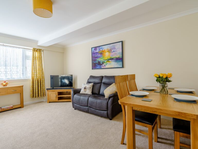 A lounge diner features a recliner sofa positioned beside a wooden dining table set for four. A television rests on a wooden stand across from the sofa. Natural light filters through a window, complemented by decorative curtains. A vase of fresh flowers adds a touch of warmth.