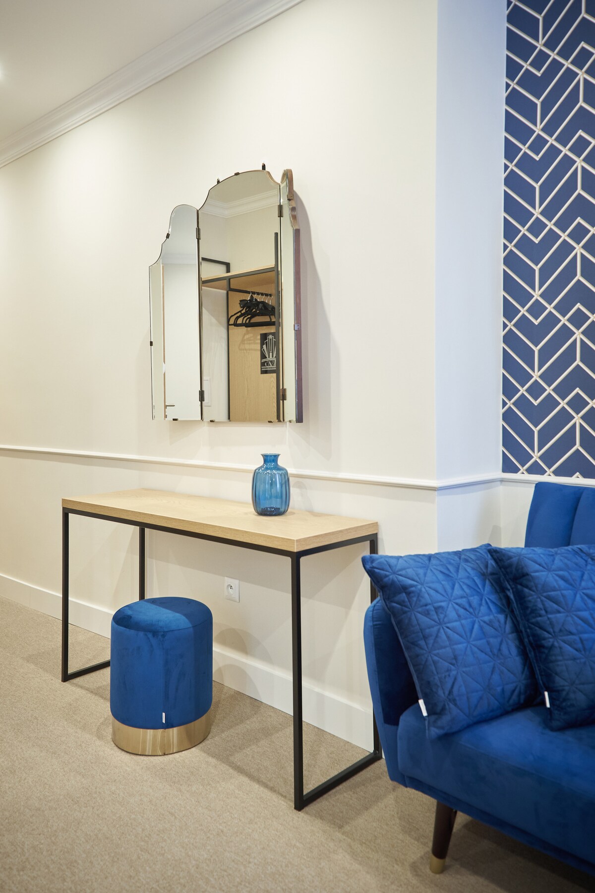 A modern entryway features a sleek console table topped with a decorative blue vase. A geometric patterned wall in blue adds visual interest. A round stool in gold and light wood is positioned nearby, alongside a plush blue couch adorned with textured cushions.