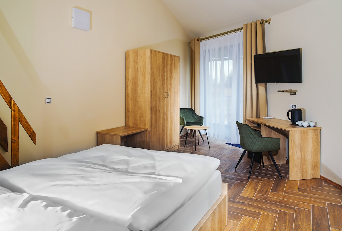A bright and airy room features a spacious bed with crisp white linens. A wooden wardrobe and a desk are visible, complemented by two green chairs. Natural light filters through sheer curtains covering a large window, and a flat-screen TV is mounted on the wall.