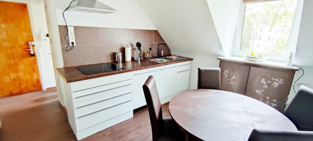 The kitchen area features modern cabinetry and a sloped ceiling. Essential appliances, including a stove and sink, are visible. A small round table with four chairs provides a dining space. Natural light enters through a window, offering a view of the outside.