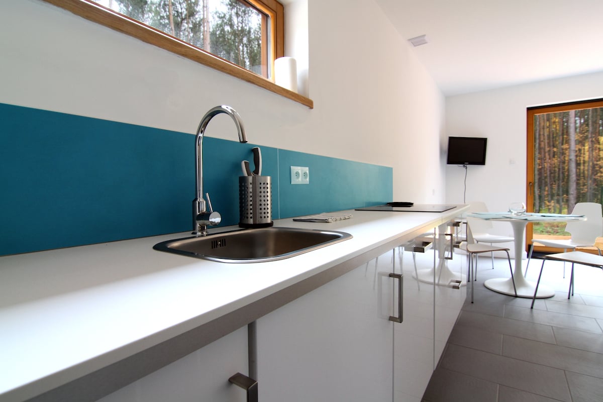 A modern kitchen is showcased, featuring a sleek white countertop and a vibrant blue backsplash. A stainless steel sink and kitchen utensils are neatly arranged. Natural light filters through a large window, illuminating the dining area with a glass table and white chairs.