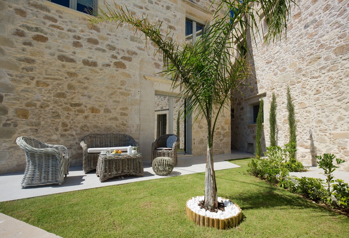 An outdoor seating area features a pair of woven sofas and a round coffee table, surrounded by well-maintained greenery. A small palm tree is centered in the garden, providing a touch of nature against the stone wall backdrop.