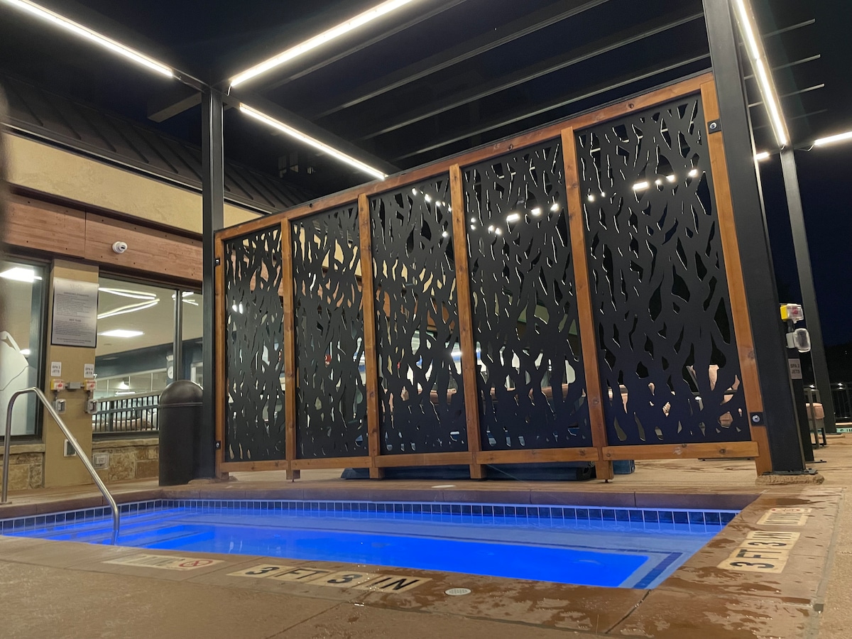 A heated outdoor pool area is illuminated at night, featuring a modern privacy screen with a leafy pattern. The pool is surrounded by clean tiles and soft blue water, with steps leading into the water and overhead lighting highlighting the inviting space.
