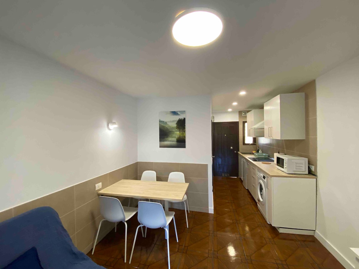 A dining area features a wooden table surrounded by four white chairs. Moderately styled cabinetry and appliances are visible in the kitchen area. A light fixture illuminates the space, enhancing the neutral color palette of the walls and flooring.