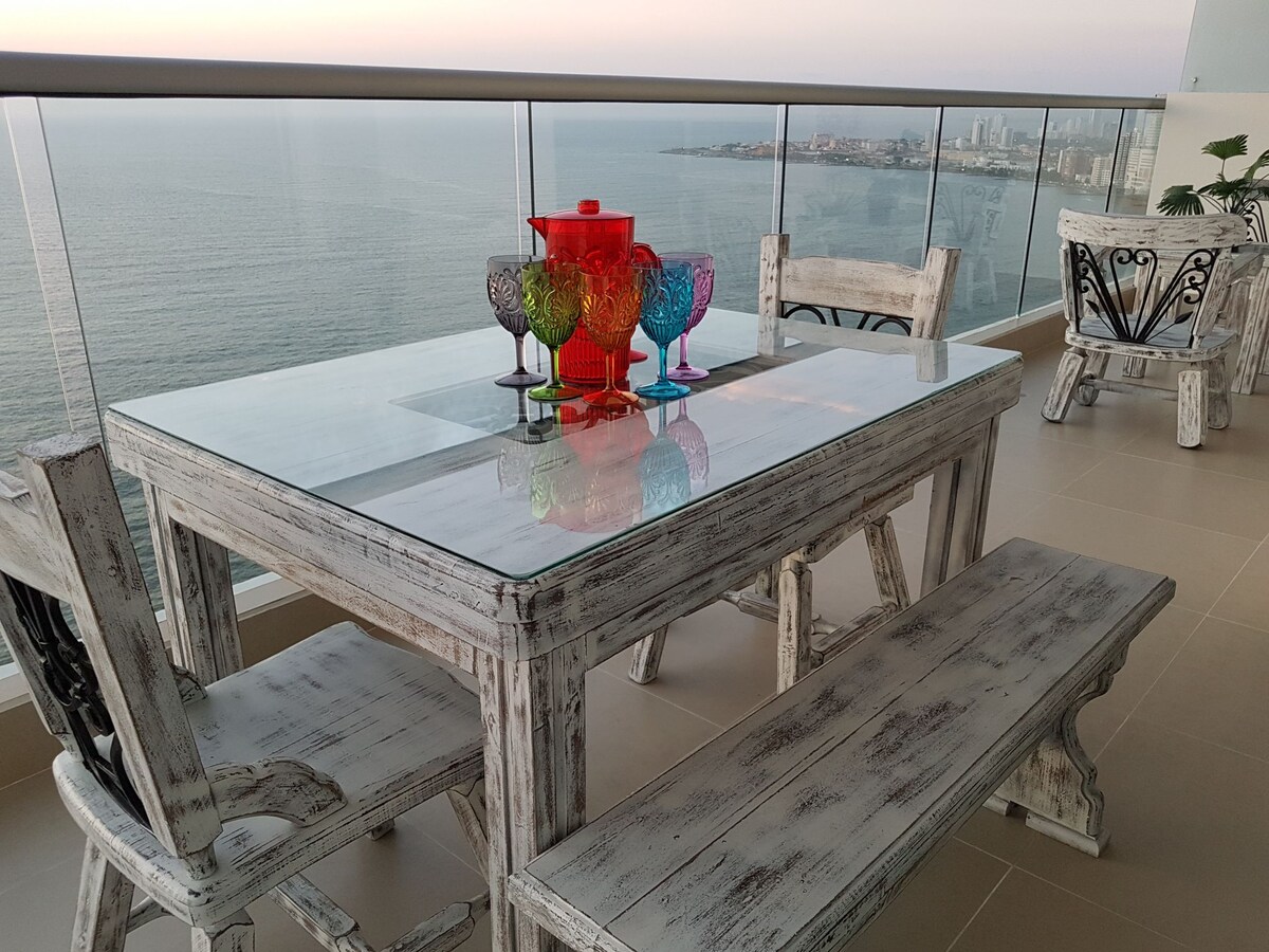 A dining area features a large glass table accompanied by rustic white wooden chairs and a bench. Brightly colored glasses and a vibrant pitcher are arranged on the table, with a serene ocean view visible through the balcony railing.