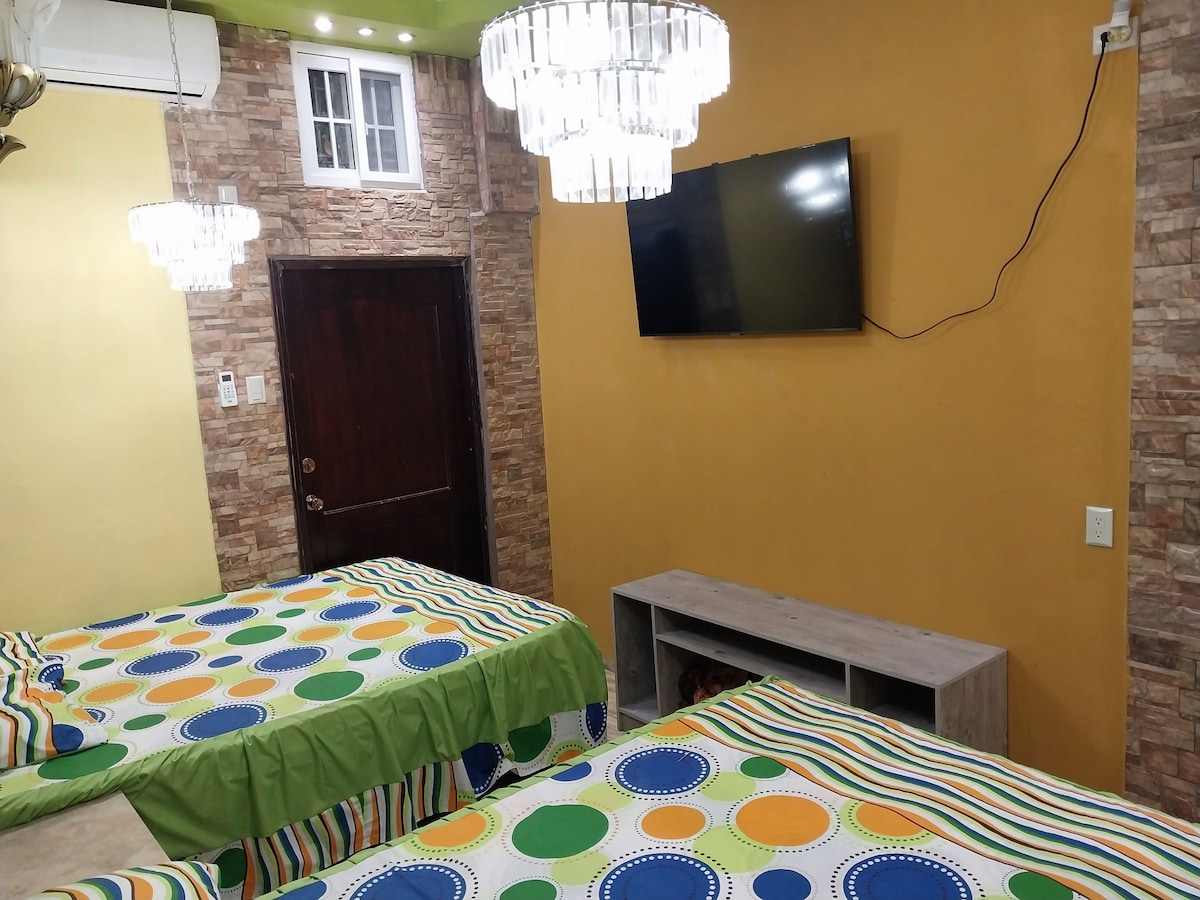 A room features two spacious beds, each adorned with colorful patterned bedding. A flat-screen television is mounted on the wall, and a wooden console table sits below it. Modern light fixtures provide illumination, and a stone accent wall complements the vibrant yellow walls.