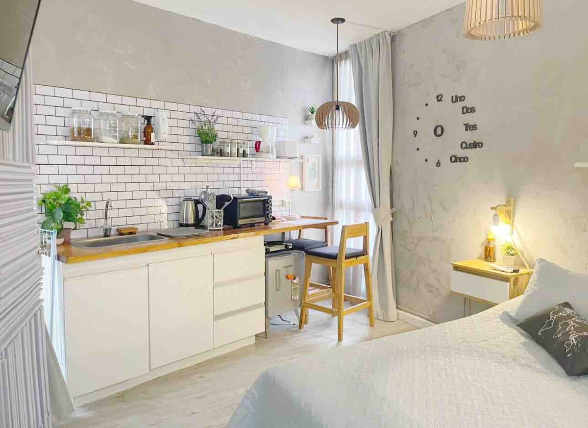 The studio features a well-equipped kitchenette with modern appliances and a wooden countertop. A dining area with a high stool is visible, while soft natural light filters through the window. The cozy sleeping area is adorned with light textures and subtle decor.
