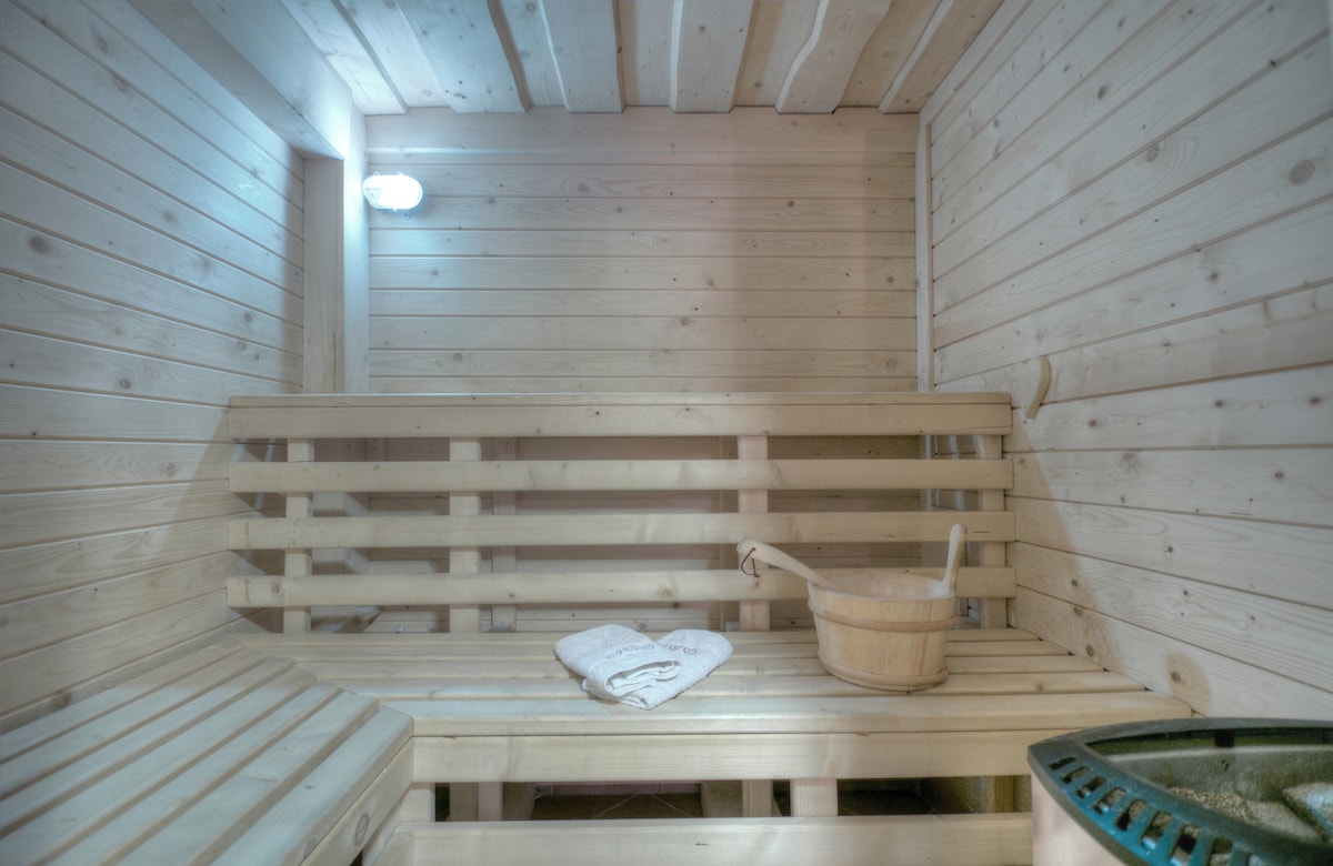 An inviting sauna space is presented with light wood paneling. Benches are arranged along the walls, and a wooden bucket is placed on the floor alongside a folded towel. Soft light illuminates the area, creating a serene atmosphere.