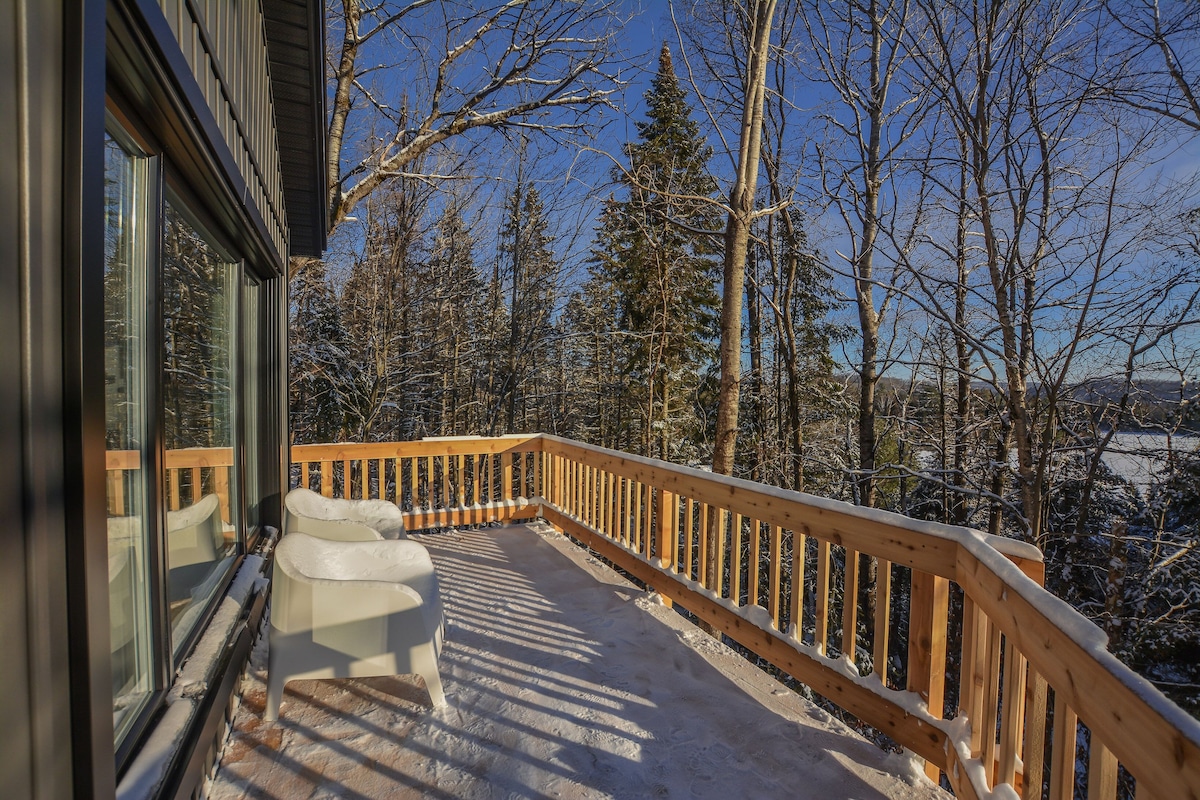 A spacious terrace is surrounded by trees and covered in snow, offering an inviting view of the winter landscape. Two white chairs are positioned on the terrace, allowing for relaxation amidst nature. Large windows of the chalet reflect the sunlight streaming through.