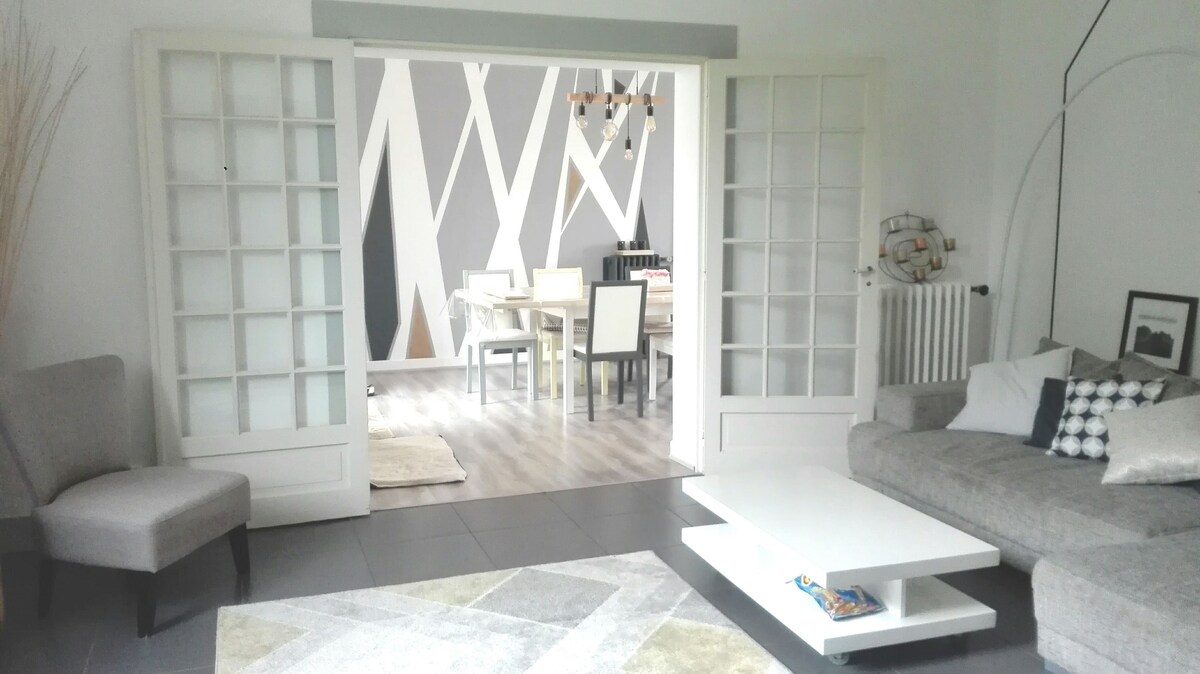 An open living space is presented, featuring a comfortable sectional sofa and a light area rug. The room is enhanced by large glass doors leading to a dining area, which is furnished with a wooden table and white chairs. Bright walls are accented with a modern geometric design.