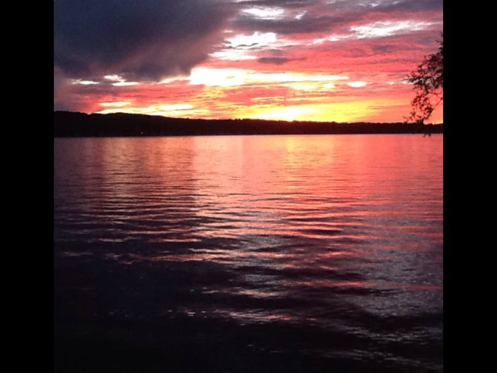 A vibrant sunset paints the sky in shades of orange, pink, and purple, reflecting off the calm waters of Canandaigua Lake. The serene landscape is framed by distant hills, creating a peaceful and picturesque view.