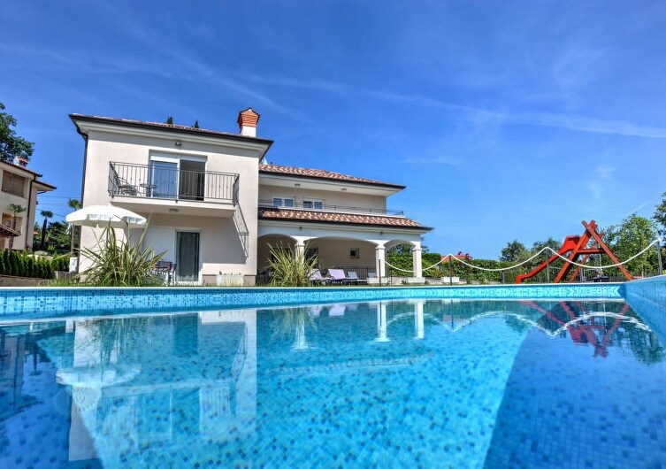The villa showcases a modern two-story design with a flat roof and balcony. A sparkling swimming pool reflects the clear blue sky, surrounded by lush greenery. Lounge chairs are positioned nearby, and a colorful children's play structure is visible on the adjacent lawn.