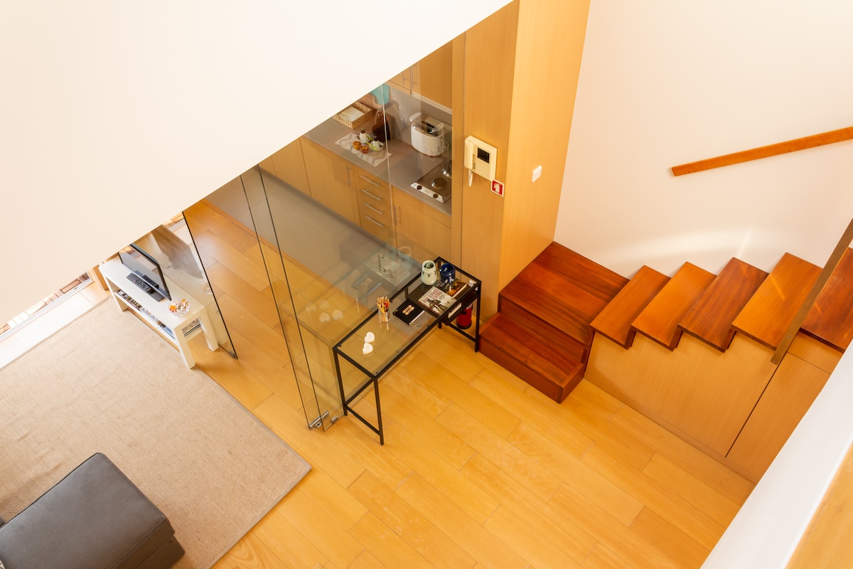 A view from above showcases the open space layout of the apartment. A glass partition separates the living area from the kitchen, while wooden stairs lead to the mezzanine level. Natural light fills the space, reflecting off the light-colored wood flooring.