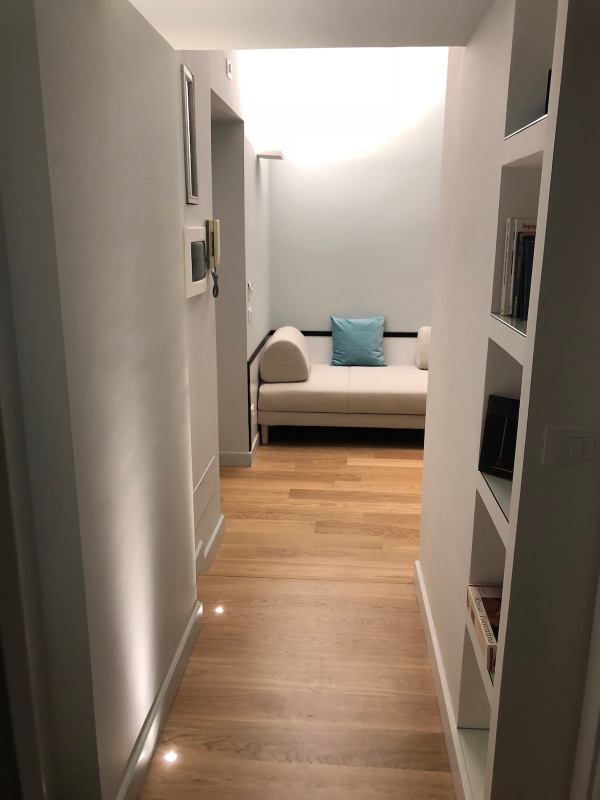 A well-lit hallway leads to a comfortable sofa adorned with a single blue cushion. Soft wood flooring extends throughout. Shelves along one wall display various books, contributing to a cozy yet modern atmosphere.